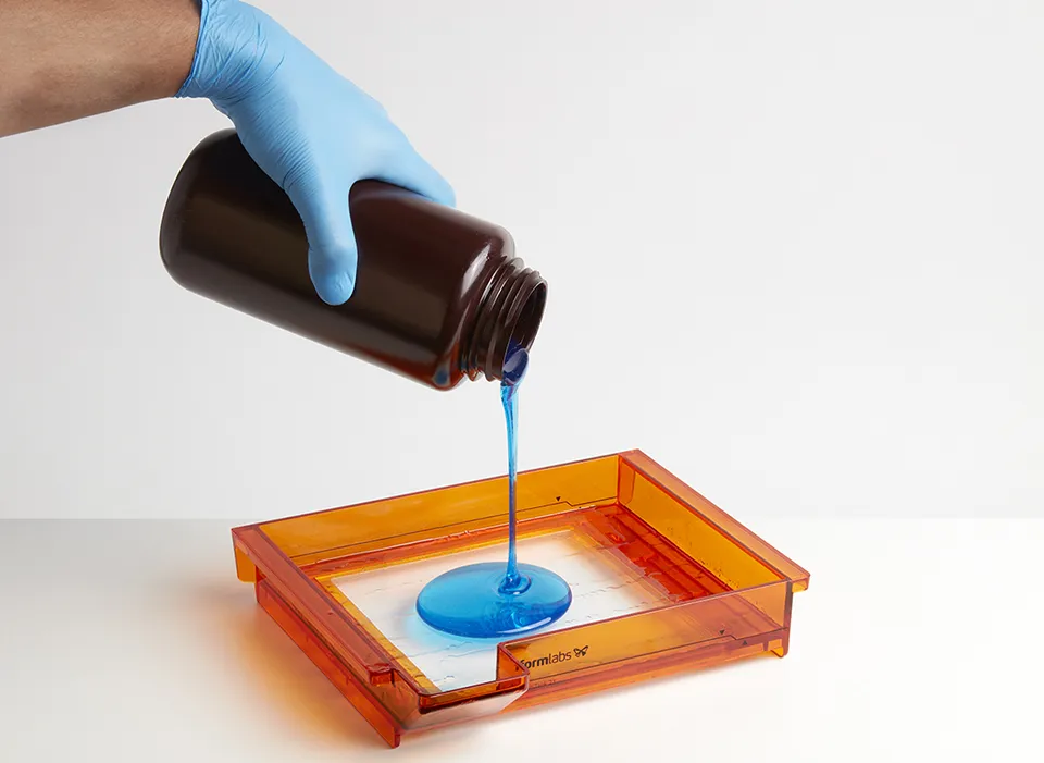 sla-3d-printer-liquid-photopolymer-resin-tank-preparation copy Mavi eldivenli bir elin, turuncu renkli şeffaf reçine tankına koyu renkli bir şişeden mavi sıvı fotopolimer reçine döktüğü anın yakından görünümü.