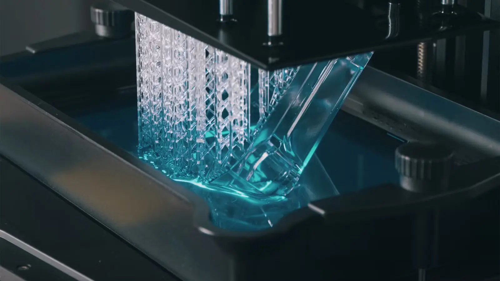 sla-3d-printing-layer-by-layer-platform-movement Mavi renkli sıvı reçine havuzunun içinden yukarı doğru yükselen, şeffaf malzemeden üretilmiş ve ince destek yapılarına sahip 3D baskı parçasının detay görünümü.